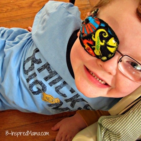 A happy smiling young child wearing eyeglasses with a handmade fabric eye patch on them. The eye patch features a colorful lizard pattern.