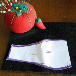 A paper template in the shape of eyeglasses' lense and temple pinned onto black felt with a pincushion with pins sitting nearby.