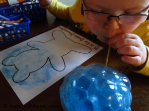 A photo of a young child blowing through a straw into a bowl to create billowing blue bubbles for a bear-themed letter B activity for preschool.
