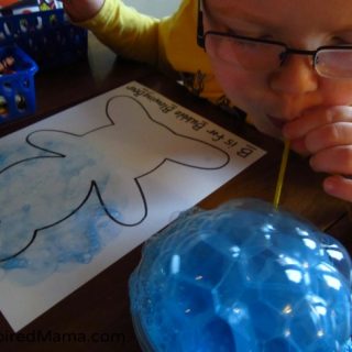 A photo of a young child blowing through a straw into a bowl to create billowing blue bubbles for a bear-themed letter B activity for preschool.