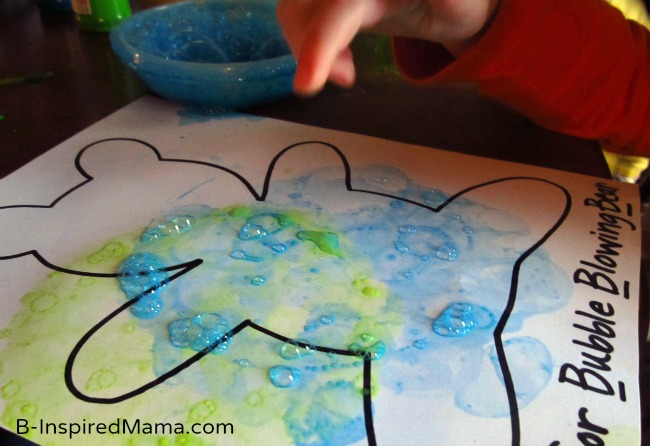 A photo of a young child poking at blue bubbles on a bear-themed letter B craft for preschool.