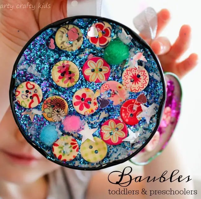 A child holds a round colorful ornament craft for preschoolers featuring beads and buttons glued inside a recycled plastic lid.