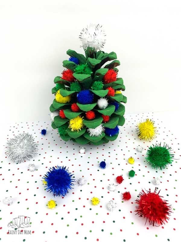 A photo of an easy preschool ornament idea made out of a pine cone painted green with colorful pom poms ticked all around it to look like a mini Christmas tree.