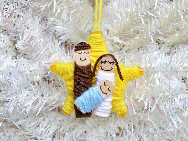 A photo of a nativity Christmas ornament craft for preschoolers featuring a star made out of craft sticks wrapped in yellow yarn topped with little craft stick nativity figures wrapped in brown, white, and light blue yarn.
