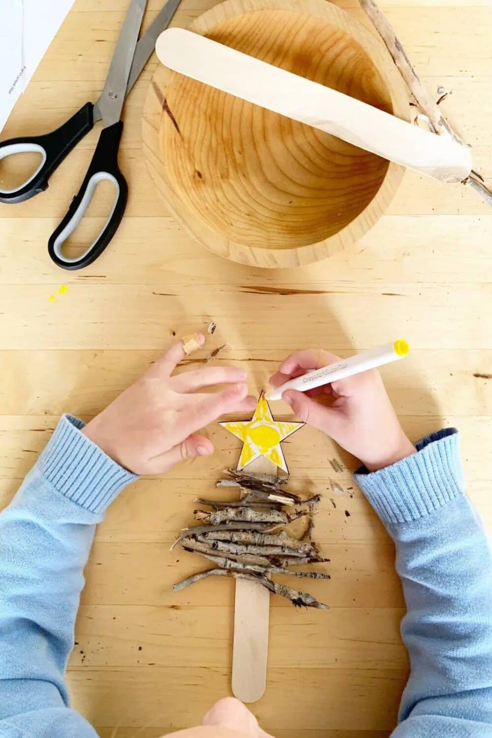 A child's hands working on a Christmas ornament craft by gluing sticks and a yellow star onto a craft stick to look like a little Christmas tree.