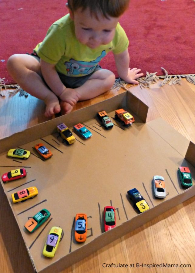 A toddler child doing a DIY cardboard parking lot number match activity with small toy cars each labeled with a different number, parking them in numbered parking spaces drawn inside the bottom edges of a cardboard box.