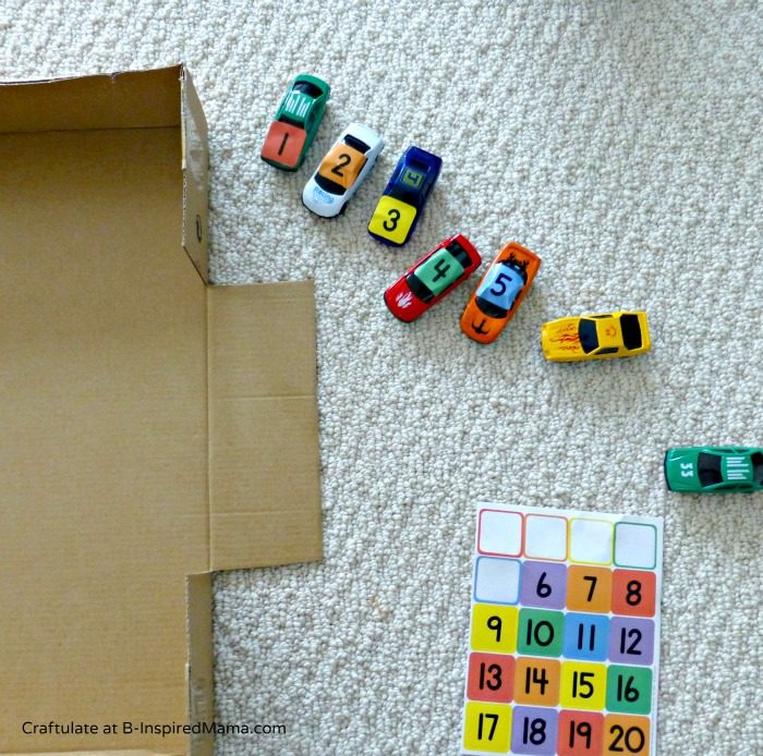 Materials for making an easy cardboard parking lot, including a large cardboard box with 3- to 4-inch walls and a ramp cut into one side. Small toy cars labeled with number stickers sit on the floor outside the box.