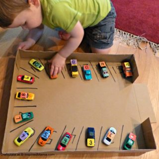 A toddler child doing a homemade cardboard parking garage number activity with small toy cars each labeled with a different number, parking them in numbered parking spaces drawn inside the bottom edges of a cardboard box.