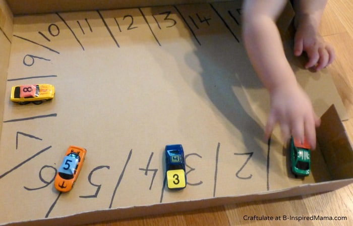 A toddler child playing with a homemade car-themed number learning activity. The activity includes a cardboard box labeled with numbered parking spaces inside and small tow cars labeled with coordinating number stickers parked in each space.