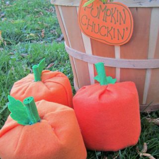 Three handmade pumpkins, made by covering toilet paper rolls with orange felt, sit in the grass alongside a tan bushel basket, with a pumpkin-shaped paper sign reading "Pumpkin Chuckin'" tied to its handle.