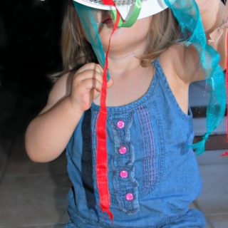 A toddler holding up and playing with a DIY toy made out of a plastic kitchen colander threaded with ribbons. The ribbons have knots tied in each end and the toddler is pulling them through the colander's holes.