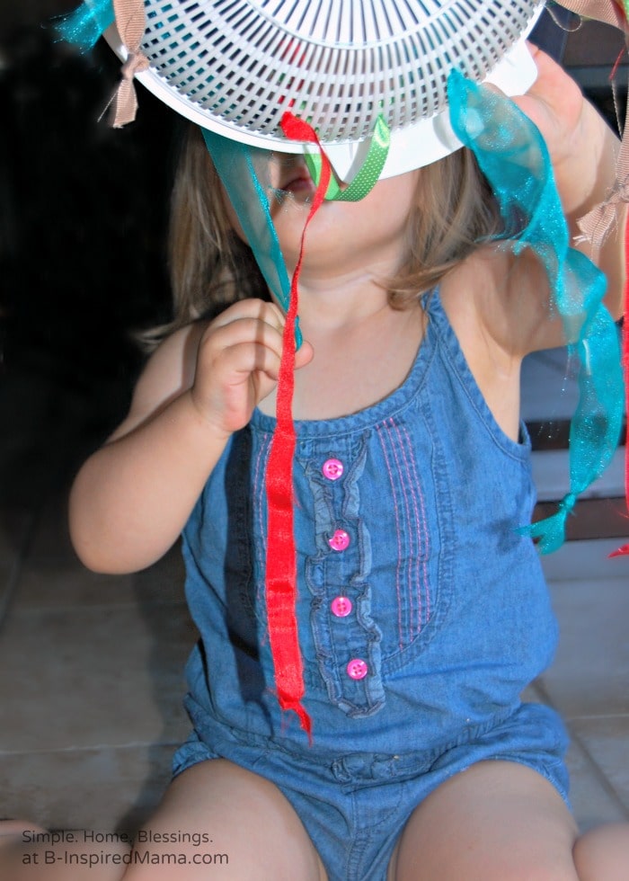 A toddler holding up and playing with a DIY ribbon pull toy made out of a plastic kitchen colander threaded with ribbons. The ribbons have knots tied in each end and the toddler is pulling them through the colander's holes.