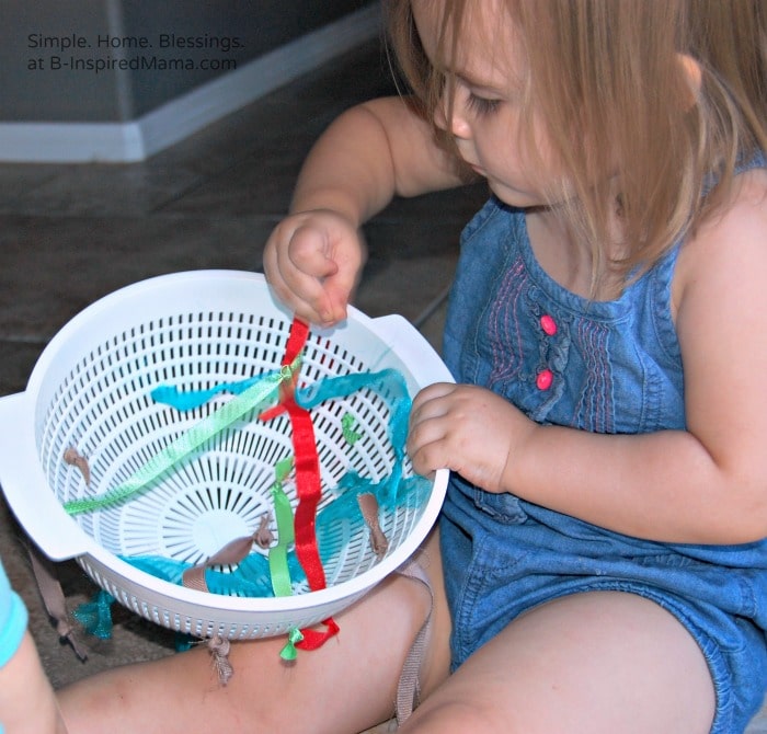 A toddler holding and playing with a DIY ribbon pull toy made out of a plastic kitchen colander threaded with ribbons. The ribbons have knots tied in each end and the toddler is pulling them through the colander's holes.