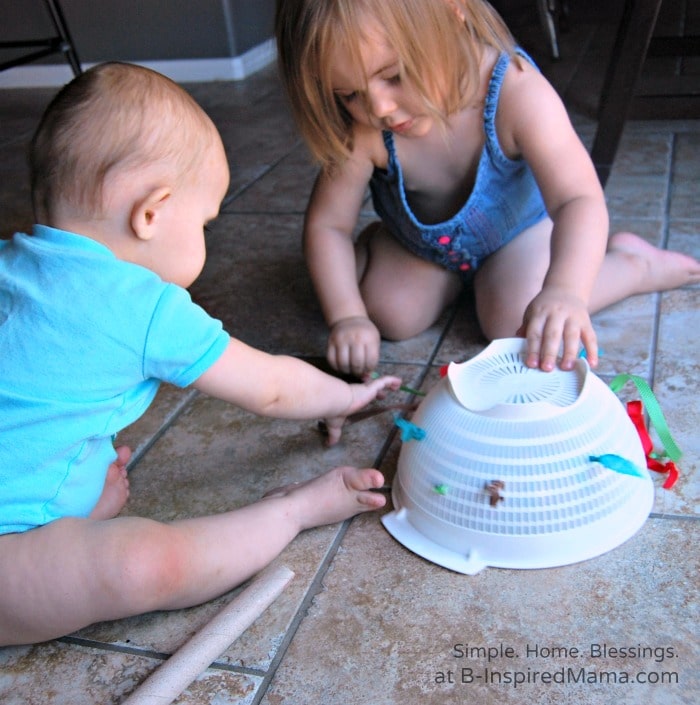 Two toddlers playing with a DIY ribbon pull toy made out of a plastic kitchen colander threaded with different colored ribbons with knots tied in the ends so they stay attached to the colander.
