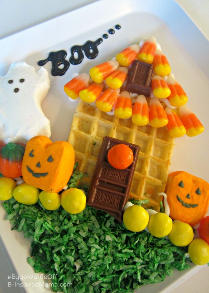 A photo of a whimsical Halloween treat consisting of a waffle cut into the shape of a Haunted house that is decorated with Halloween candy including candy corn roof tiles, chocolate bar door and window, yellow and orange M&Ms, green-dyed shredded coconut grass, and Jack-O-Lantern and ghost-shaped marshmallow Peeps.