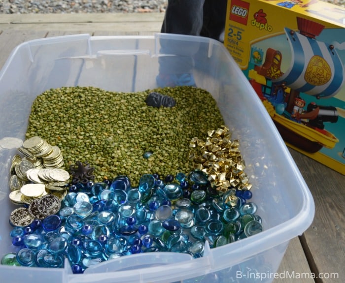 A photo of the supplies needed for LEGO Duplo Pirate Sensory Play, including a plastic bin filled with dried split peas, plastic gold coins, and golden and light blue plastic vase filler along with the boxed LEGO Duplo Pirate set.