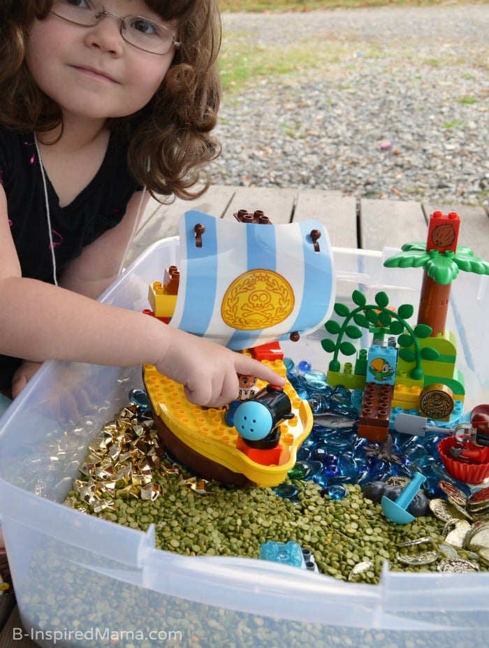 A photo of a little girl playing with a LEGO Duplo Pirate set inside a sensory bin full of dried split peas and plastic golden coins.