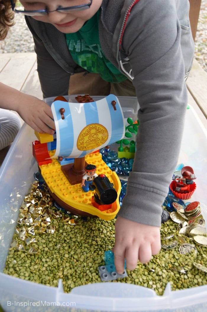 A photo of a boy playing with a LEGO Duplo Pirate set inside a sensory bin full of dried split peas and plastic golden coins.
