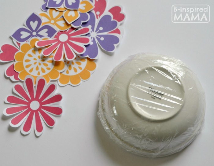 A photo of a step in the process of making a Mothers Day Flower Craft Bowl, featuring an upside-down bowl covered in plastic wrap alongside cut-out paper flowers.