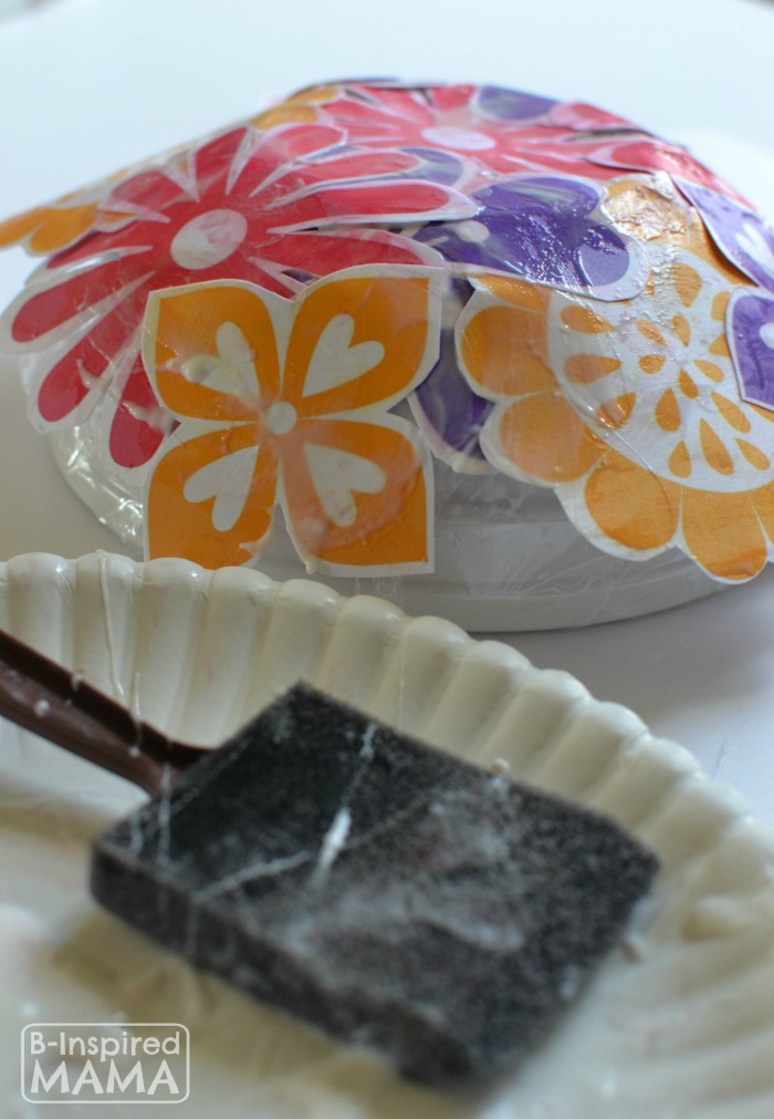 A photo of a step in the process of making a Mothers Day Flower Craft Bowl, featuring an upside-down bowl covered in plastic wrap and overlapping glue-covered paper flowers.