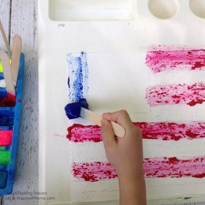 A photo of a child's hand painting an American flag with blue ice paint on a craft stick.