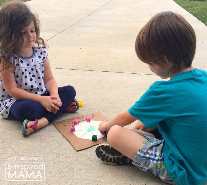 A photo of two young children playing a homemade Shisima Math Game — a math game from Kenya. The game uses a white paper octagon shape glued onto a piece of cardboard and math counters for tokens. 
