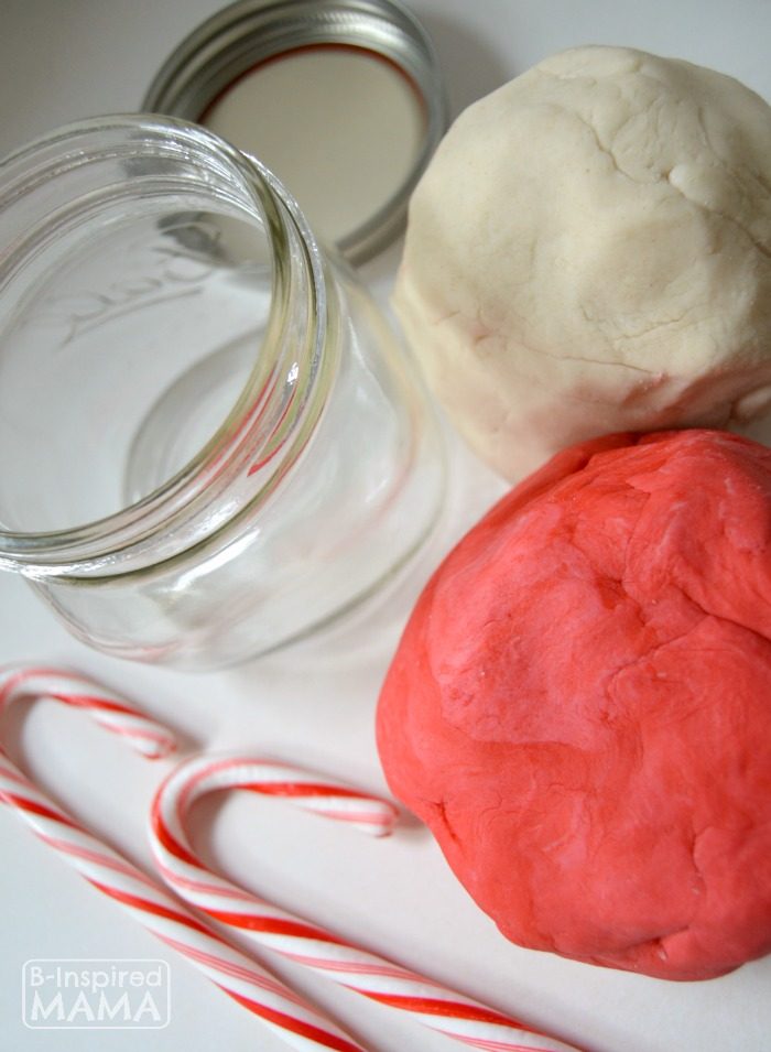 A photo of a step in the process of making DIY Christmas Playdough Jar gifts using an easy Candy Cane Peppermint Playdough recipe. The photo features balls of red and white homemade playdough alongside an open mason jar and candy canes.