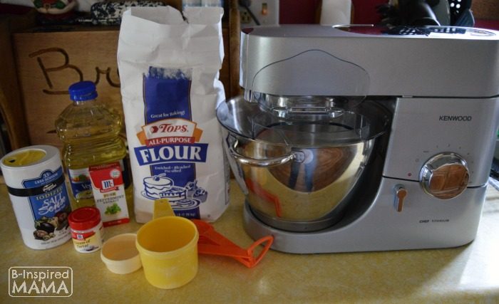 A photo of the ingredients for homemade Candy Cane Peppermint Playdough, including salt, cream of tartar, vegetable oil, all purpose flour, peppermint extract, and water, alongside a kitchen stand mixer.