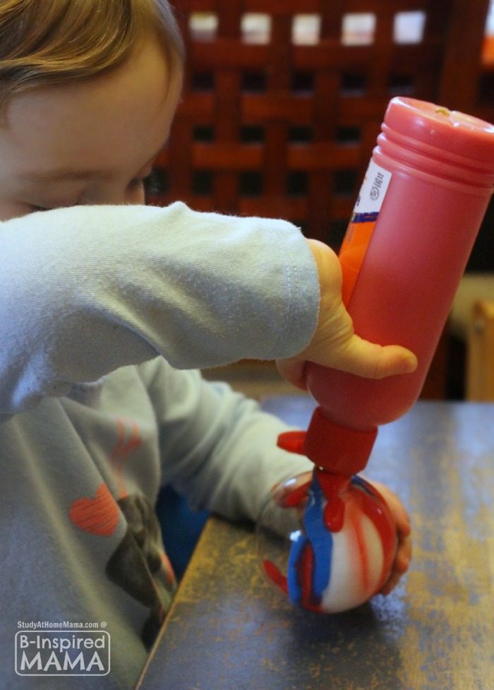A young child holds a bottle of red paint upside down, squirting paint inside a clear plastic ball ornament to make a homemade pour painted Christmas ornament.