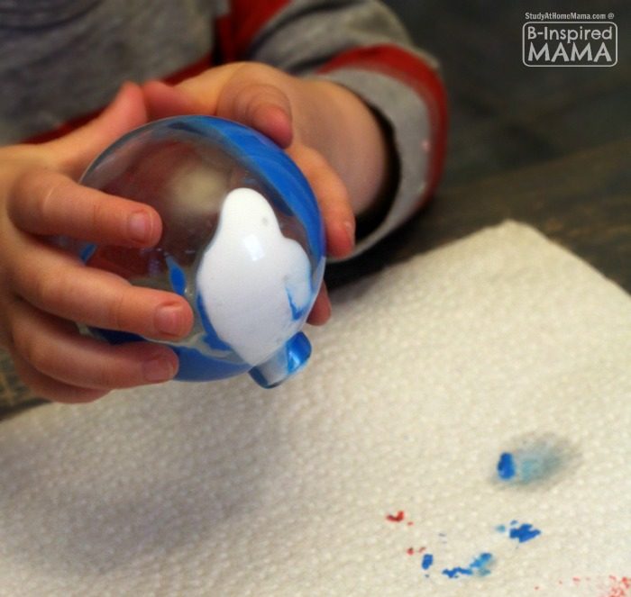 Child's hands hold clear plastic ball ornament upside down to allow blue and white paint that is inside the ornament to drip out onto paper towel.