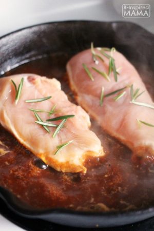 A photo of one of the steps for the recipe for Easy Skillet Rosemary Chicken and Tomatoes. The photo includes raw chicken breasts reasoned with fresh rosemary leaves inside a cast iron skillet of simmering tomato sauce.