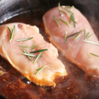 A photo of one of the steps for the recipe for Easy Skillet Rosemary Chicken and Tomatoes. The photo includes raw chicken breasts reasoned with fresh rosemary leaves inside a cast iron skillet of simmering tomato sauce.