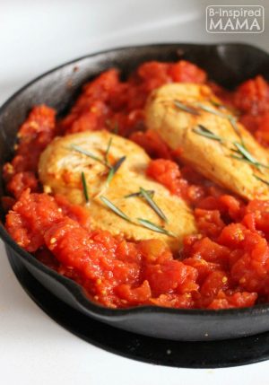 A photo of one of the steps for the recipe for Easy Skillet Rosemary Chicken and Tomatoes. The photo includes browned chicken breasts reasoned with fresh rosemary leaves inside a cast iron skillet of a thick tomato sauce.