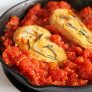 A photo of one of the steps for the recipe for Easy Skillet Rosemary Chicken and Tomatoes. The photo includes browned chicken breasts reasoned with fresh rosemary leaves inside a cast iron skillet of a thick tomato sauce.