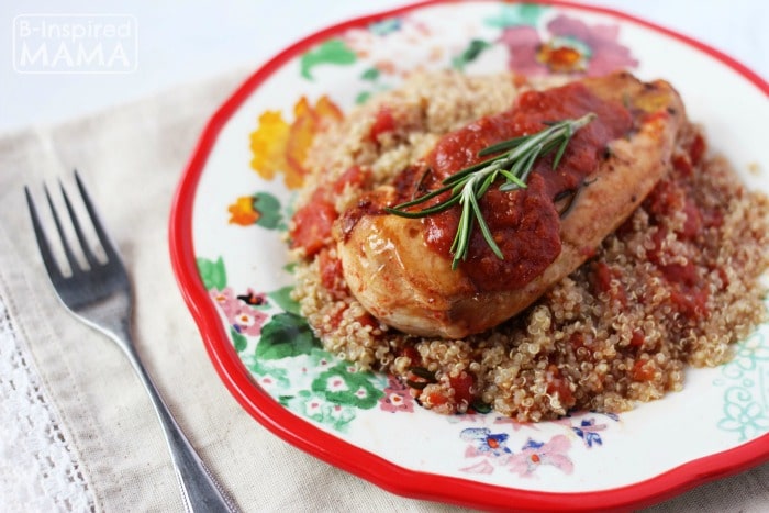 A photo of a recipe for Rosemary Chicken with Tomatoes featuring a chicken breast cooked in diced tomatoes served on top of quinoa and seasoned with a rich tomato sauce and fresh rosemary sprigs.