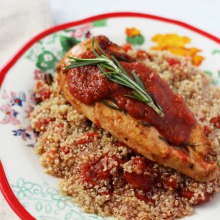 A photo of a recipe for Rosemary Chicken with Tomatoes featuring a chicken breast cooked in diced tomatoes served on top of quinoa and seasoned with a rich tomato sauce and fresh rosemary sprigs.