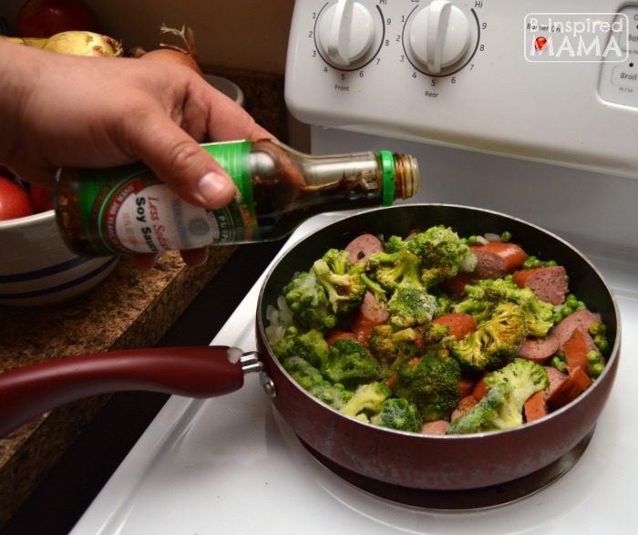 A photo of a step in the process of making a quick Smoked Sausage and Rice Skillet Recipe, including a hand pouring from a bottle of soy sauce into a frying pan filled with sliced smoked sausage and broccoli.