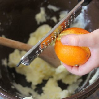 A photo of hands using a kitchen zester tool to zest an orange over a large mixing bowl.