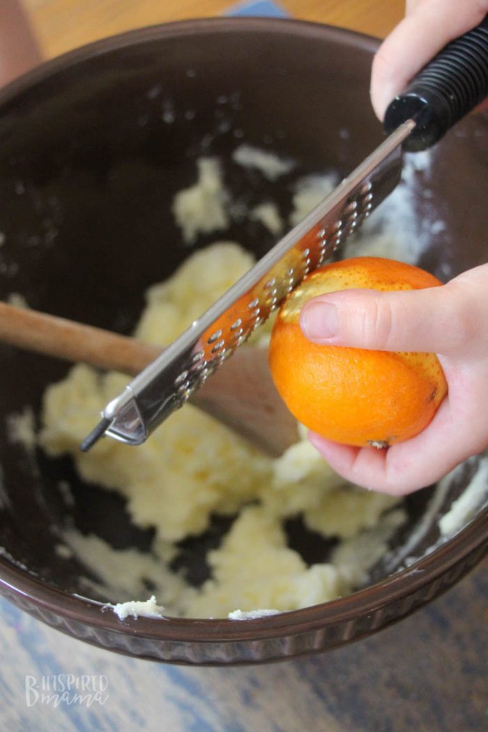 A photo of hands using a kitchen zester tool to zest an orange over a large mixing bowl.