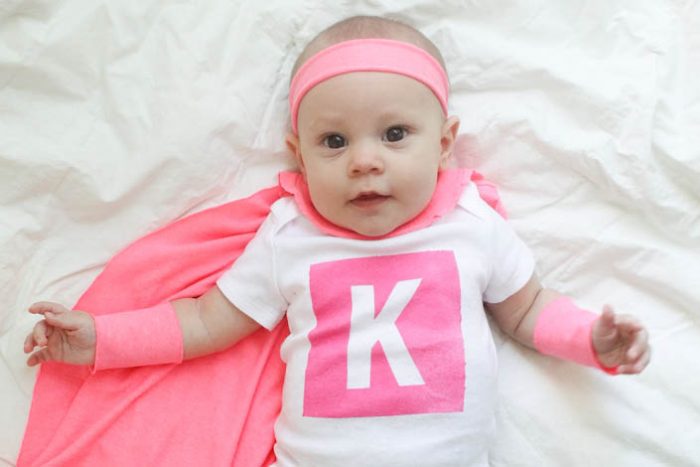 A photo of a smiling baby wearing a DIY superhero costume consisting of a white onesie with a pink letter K on it, pink armbands, a pink headband, and a pink cape. 