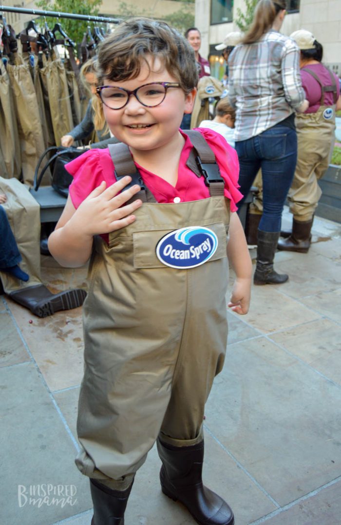 A smiling child wearing hip waders with an Ocean Spray logo on the front of them. Others are trying on hip waders in the background.