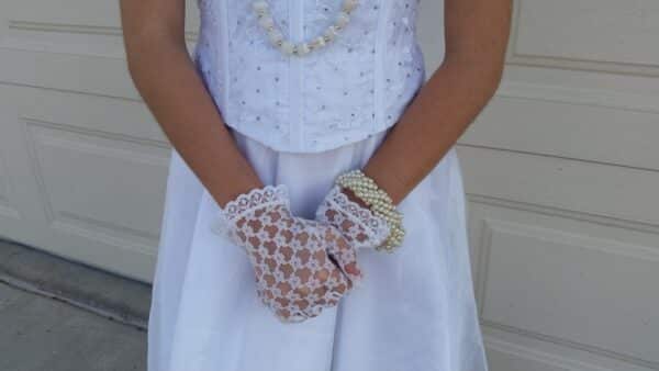 A photo of a girl's torso with her hands clasped in front of her waist. She is wearing a DIY bride Halloween costume that includes lace gloves, pearl necklace and bracelet, a long white skirt, and white top.