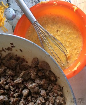 Browned and drained loose breakfast sausage in a pan alongside a mixing bowl with a mixture of eggs and shredded cheese.