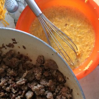 Browned and drained loose breakfast sausage in a pan alongside a mixing bowl with a mixture of eggs and shredded cheese.