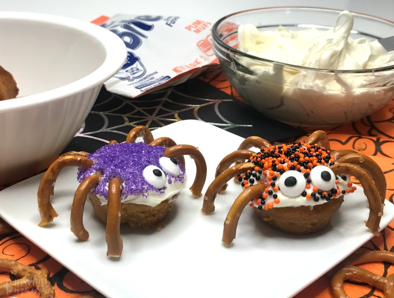 A photo of 2 small Spider Bites, a fun Halloween snack for kids consisting of Little Bites Pumpkin Muffins decorated with white frosting, Halloween sprinkles, candy eyeballs, and pretzel spider legs.