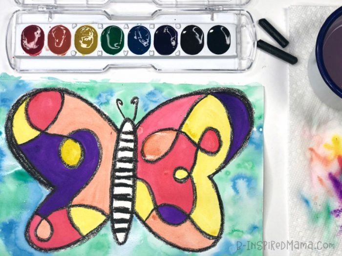 A photo of a colorful kids watercolor butterfly painting in progress, along with some kids art supplies including watercolor paints and black crayons.