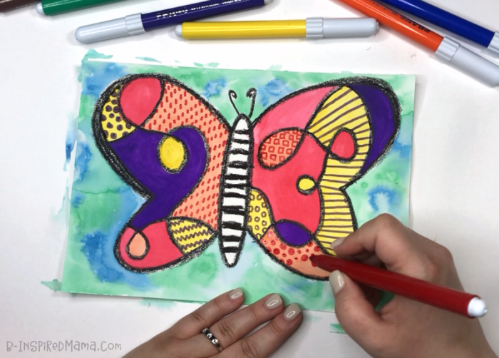 A photo of a hand using markers to draw patterns inside the wings of a kids watercolor butterfly painting.