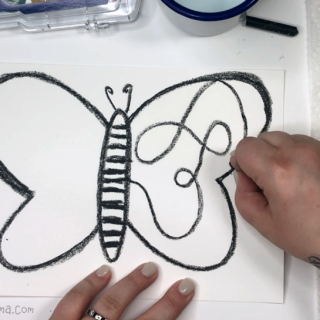 A photo of a hand using black crayon to draw a large butterfly, with swirly lines inside the wings, on a white piece of paper in preparation for a watercolor butterfly painting.
