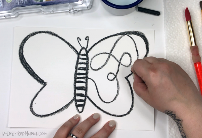 A photo of a hand using black crayon to draw a large butterfly, with swirly lines inside the wings, on a white piece of paper in preparation for a watercolor butterfly painting.