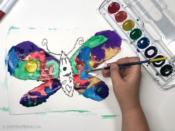 A photo of a child's hand painting a colorful watercolor butterfly painting.